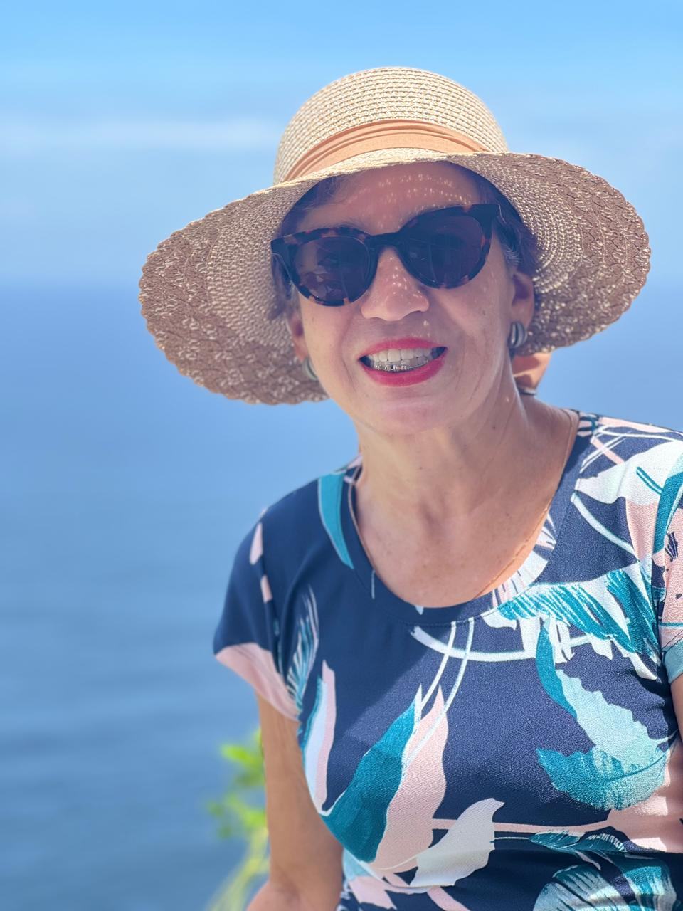 Retrato de Graça Piccolo sorrindo, de chapéu de palha, diante do mar